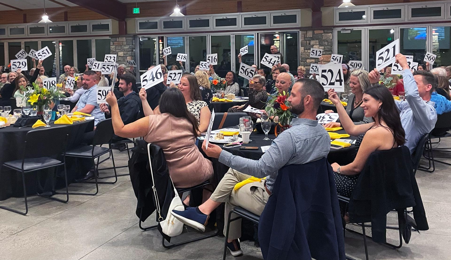 A room with people seated at round tables. Many have their hands raised, bidding on an auction item