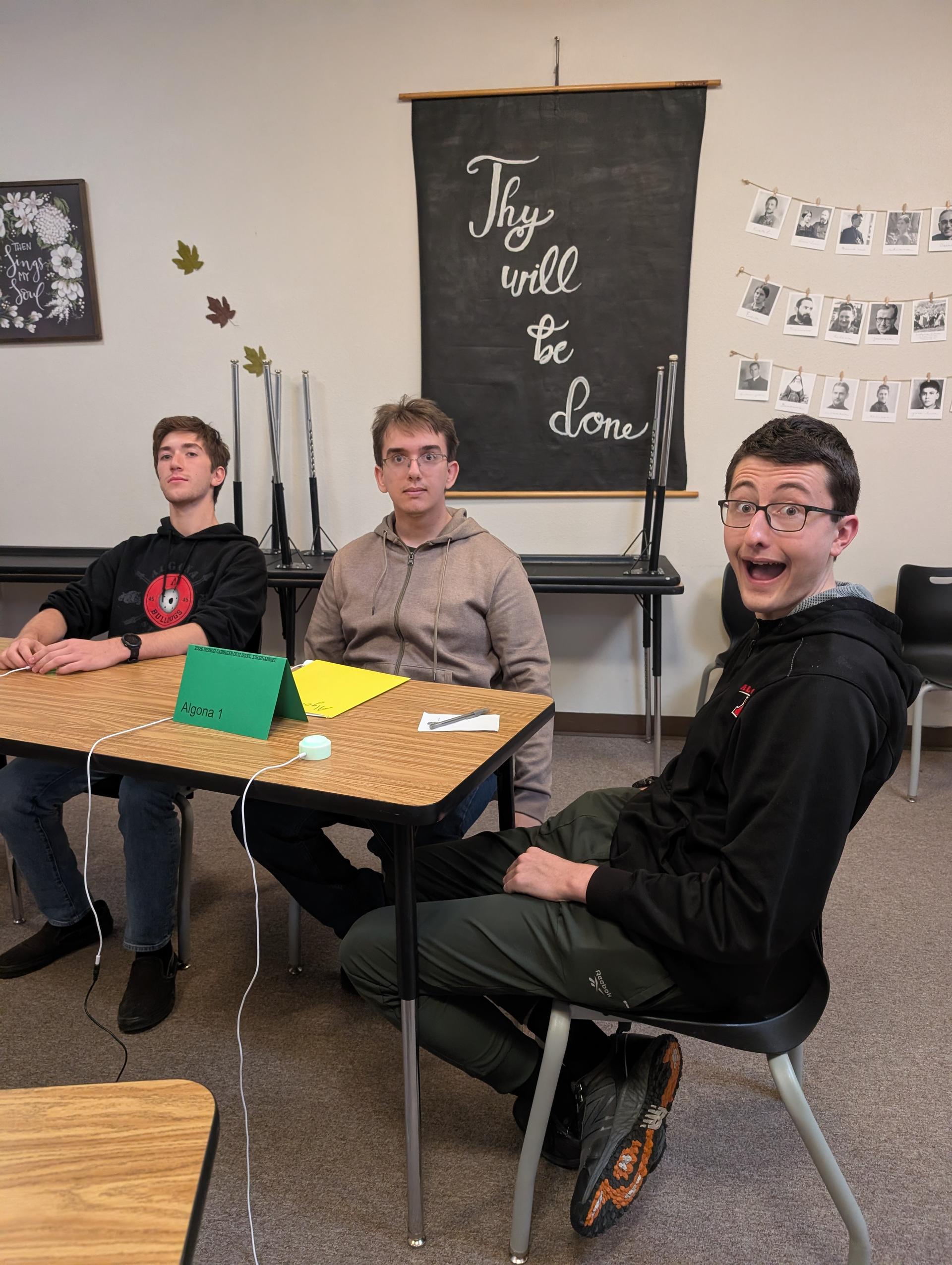Three students seated at a desk in a classroom, with one holding a small green folder and another wearing glasses.