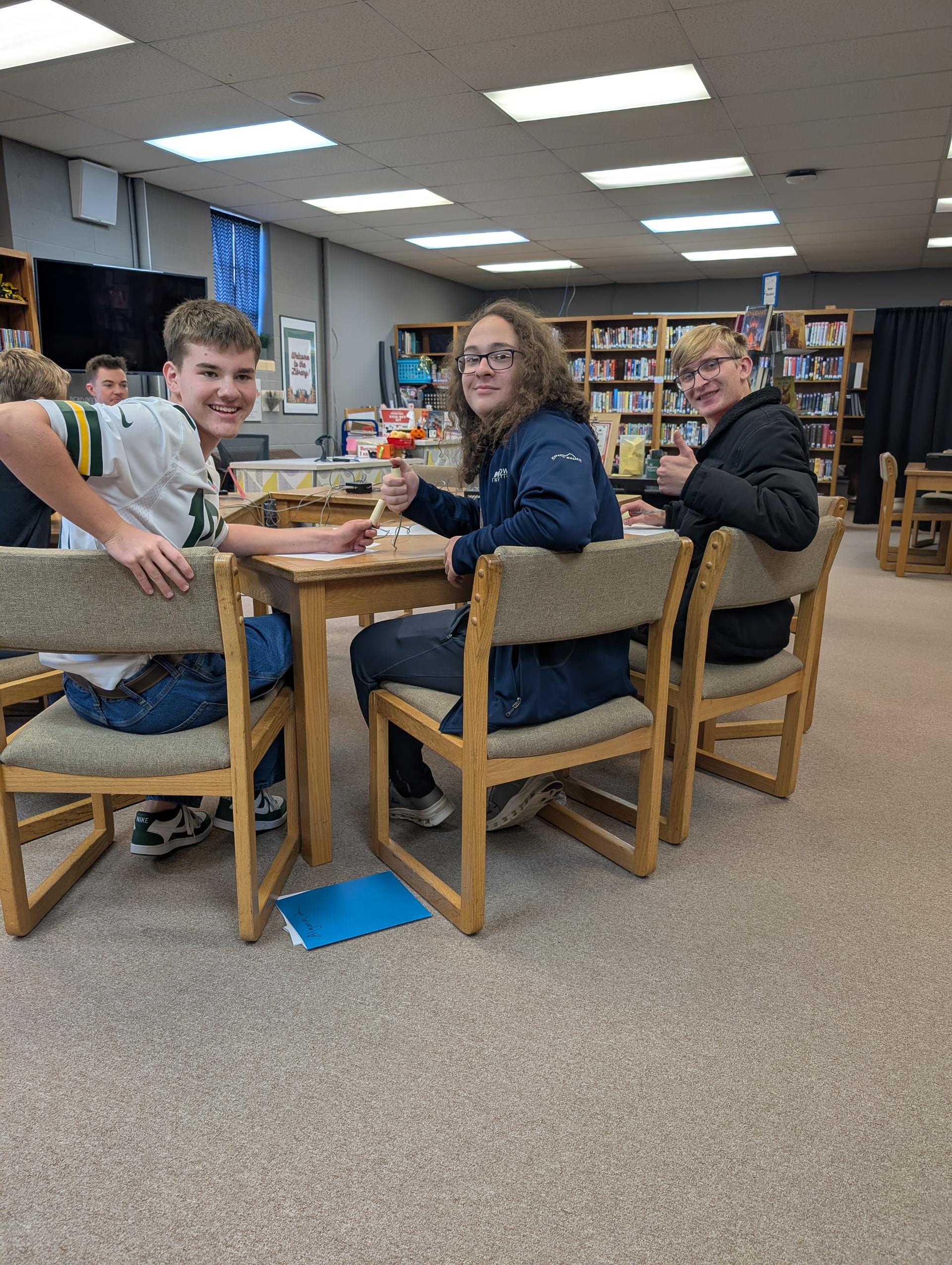 Three people at a table in a library, two of them with food, one of them holding a drink.