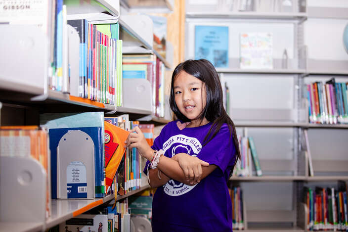 A student smiles to the camera as they pull a book out of a library shelf. 