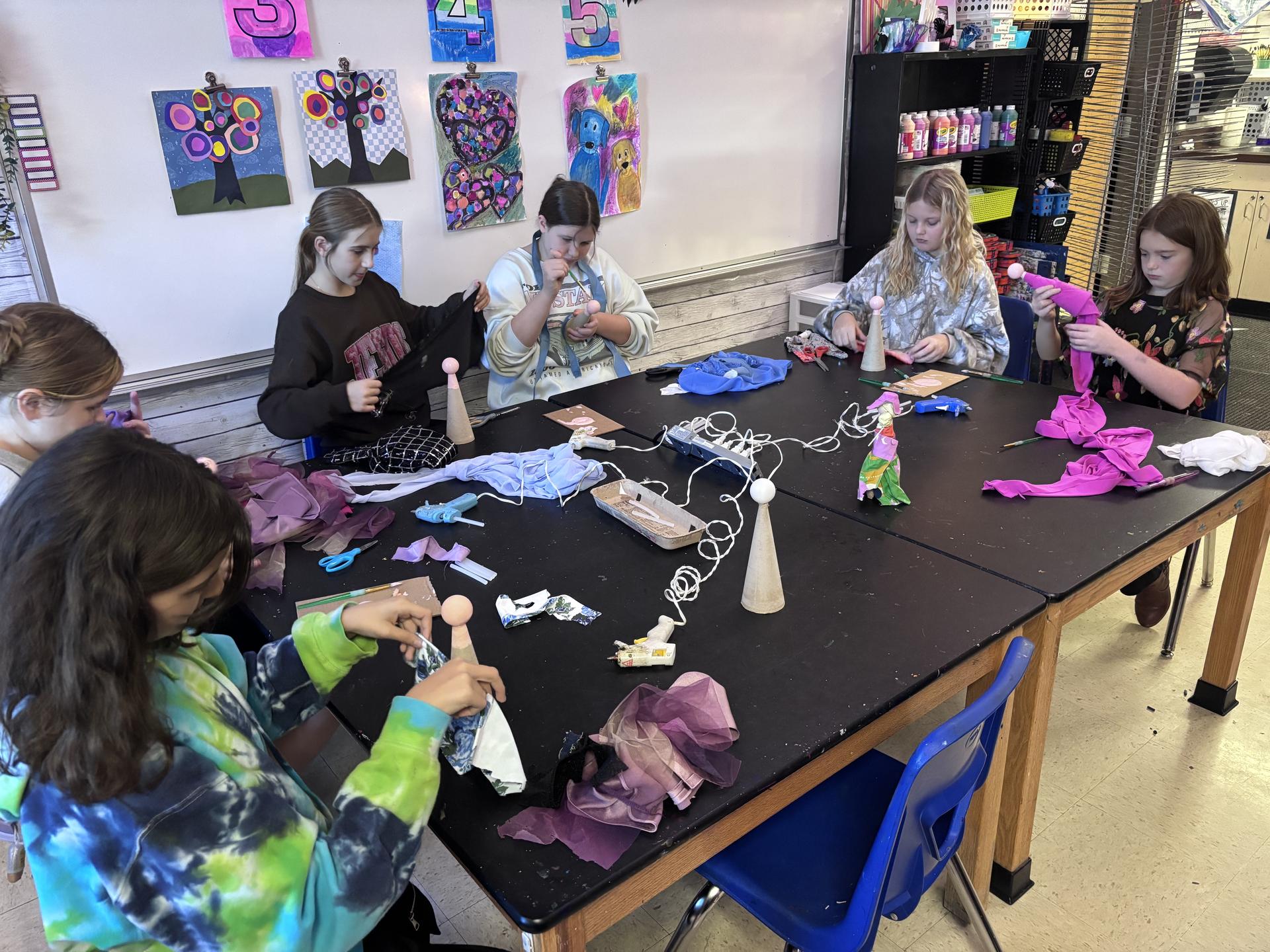 A group of female students sitting at a black table.  They are working on fabrics projects, the fabrics are of different colors. 