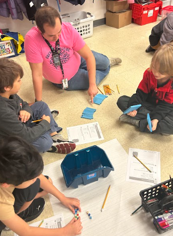 An adult guides students who have spread manipulatives and worksheets across the classroom floor
