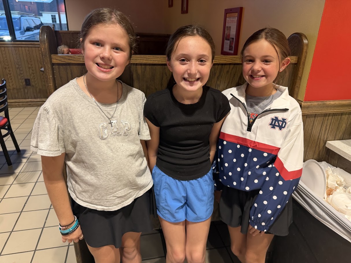 Three girls stand close together inside a restaurant with tiled flooring and booth seating. One wears a white hoodie, another a gray shirt, and the third a black top.
