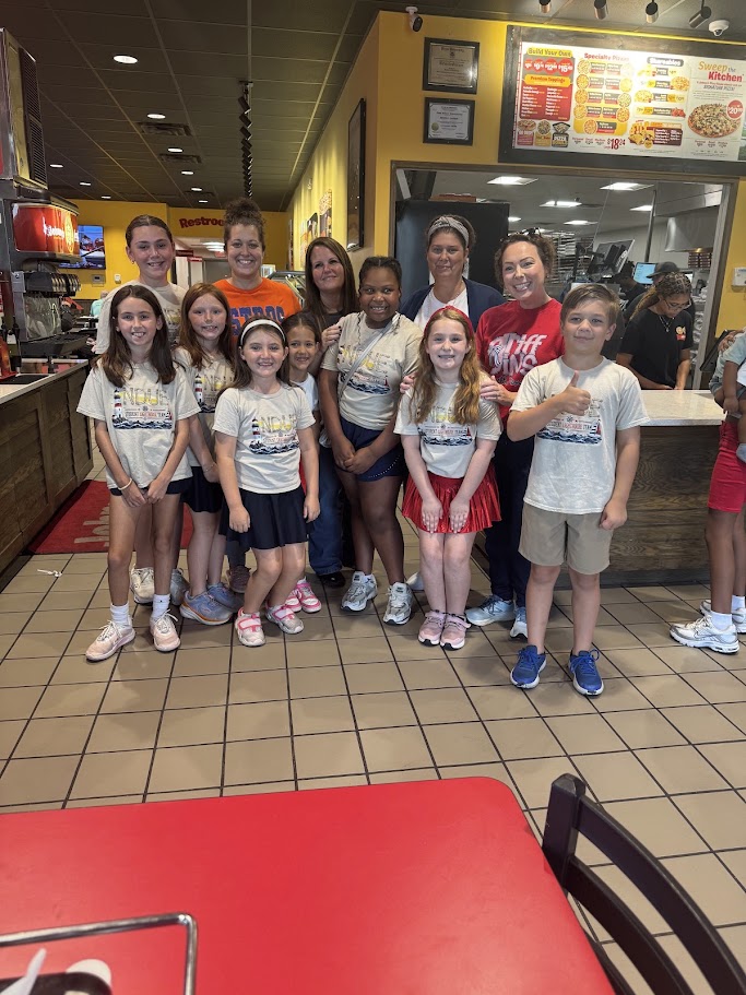 A group of children and adults stand in a restaurant with tiled floors, wearing matching shirts.