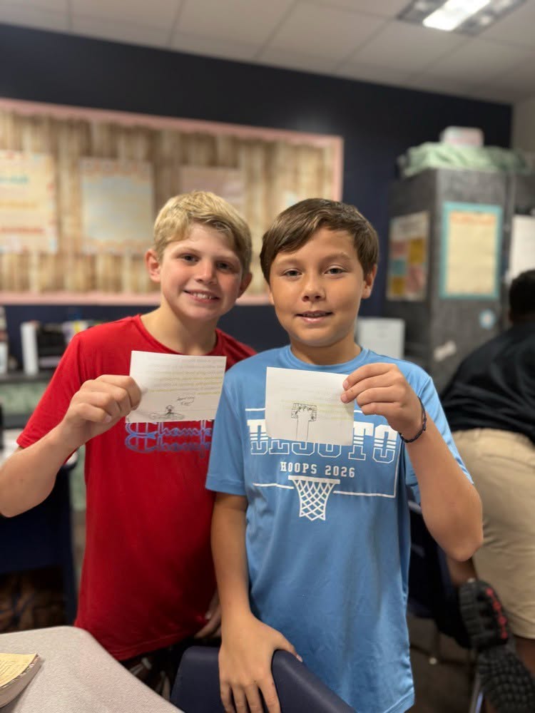Two boys in a classroom, one in red and one in blue, holding up a piece of paper.