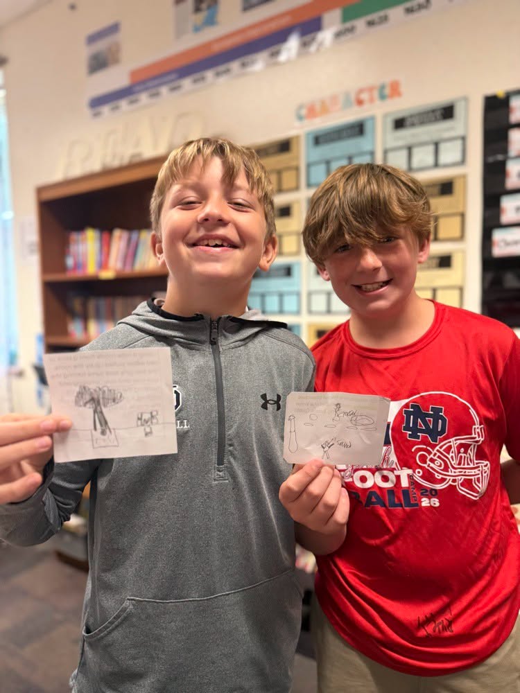 Two boys in a classroom hold drawings. One wears a gray hoodie, the other a red shirt. Behind them is a bookshelf and a bulletin board.