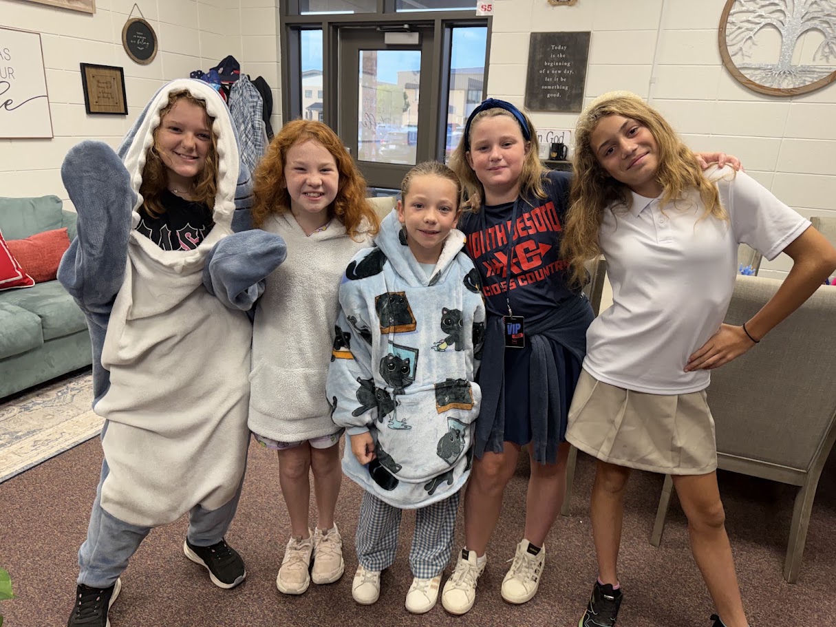 Five young girls in a room with a couch and window. Two wear white shirts, two in hoodies, one in a shark costume.