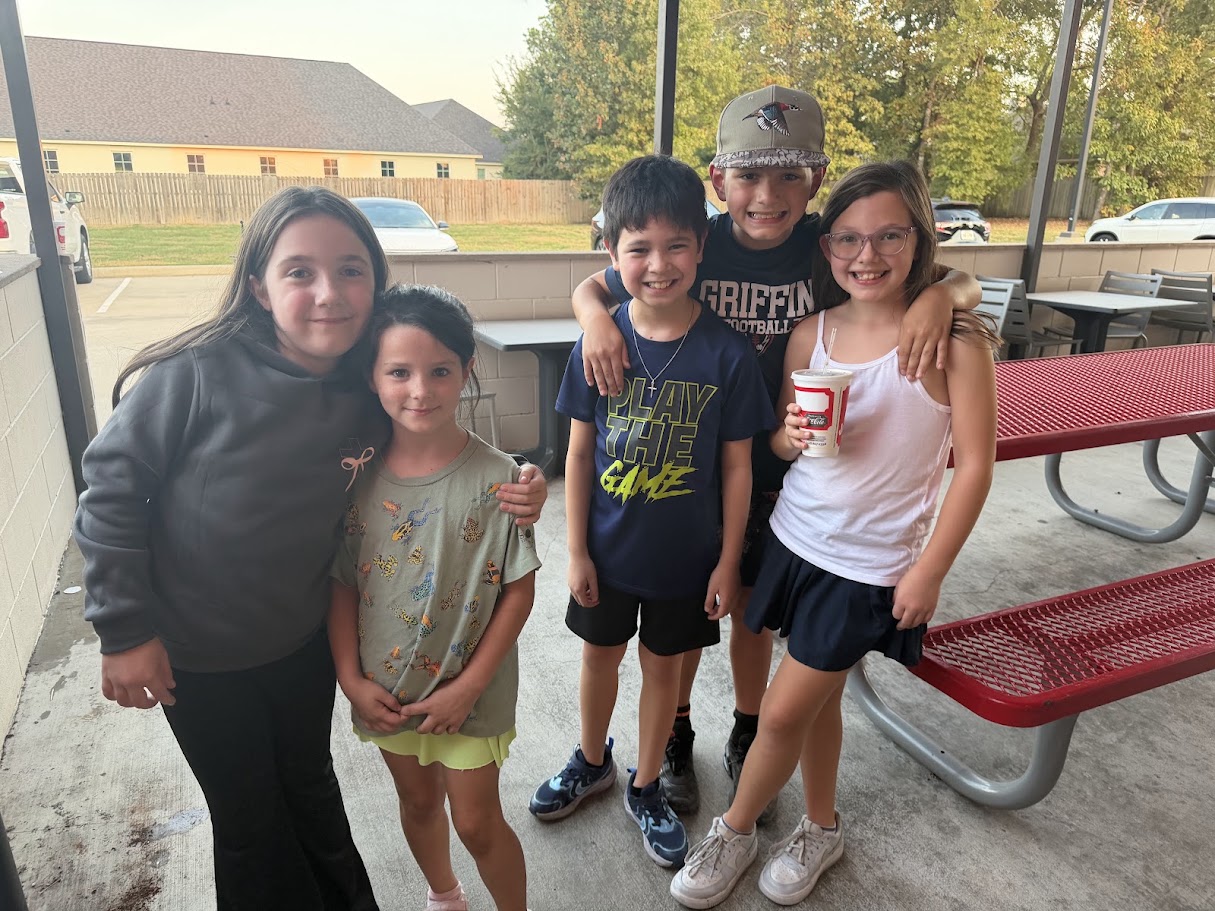 Five young people stand together at a picnic table. A woman in a gray jacket holds a young girl in a pink shirt.