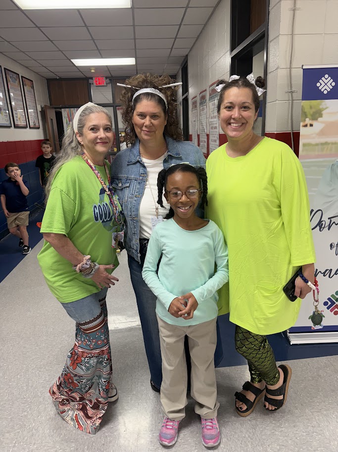 Four women in green, yellow, and blue shirts, with one in glasses, standing in a hallway.