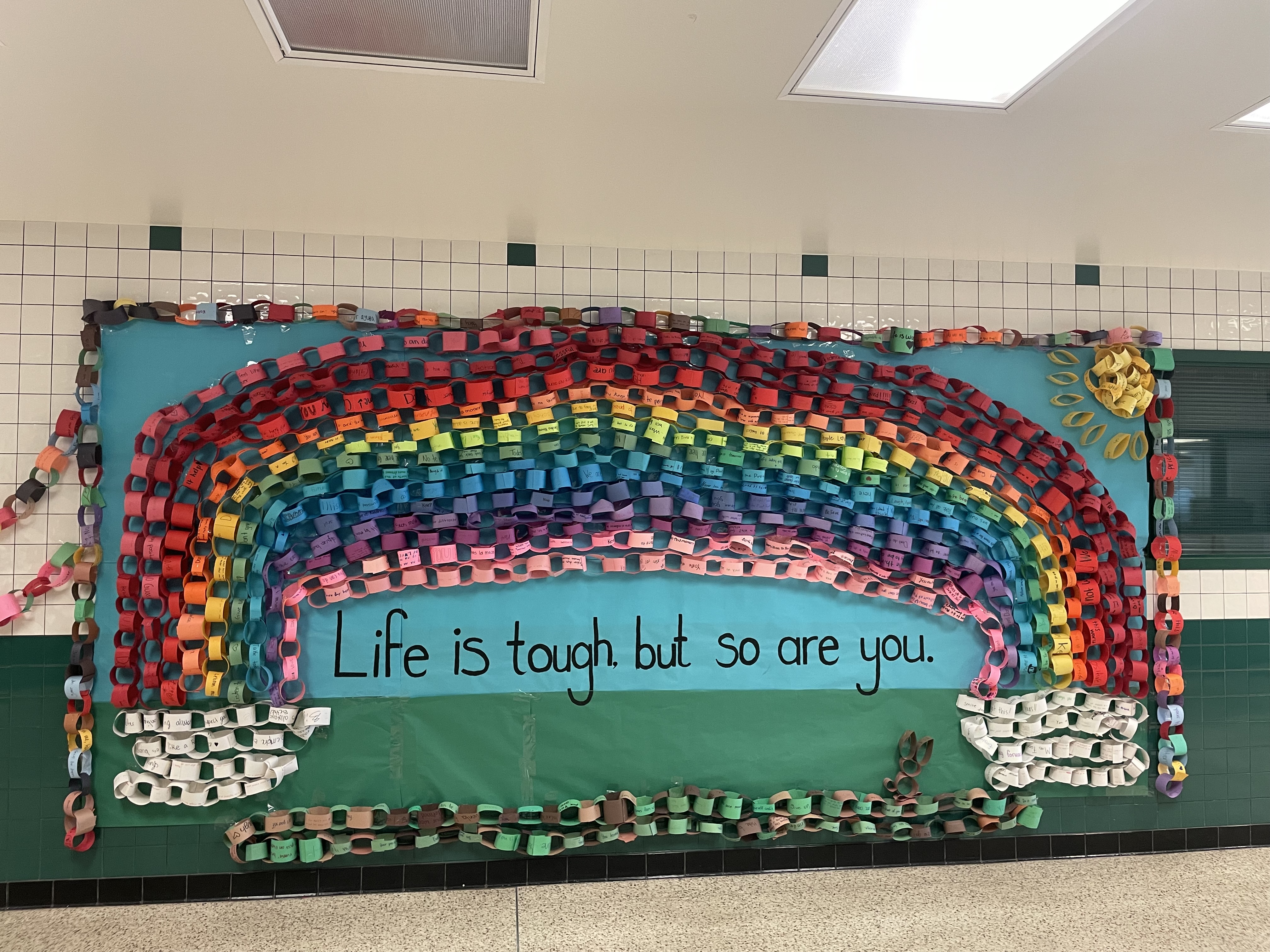 A bulletin board in a hallway with a rainbow-shaped decoration and the words 