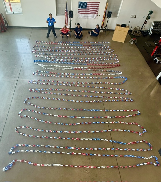 A room with a flag, a podium, and people. Long red, white, and blue strips are arranged on the floor.
