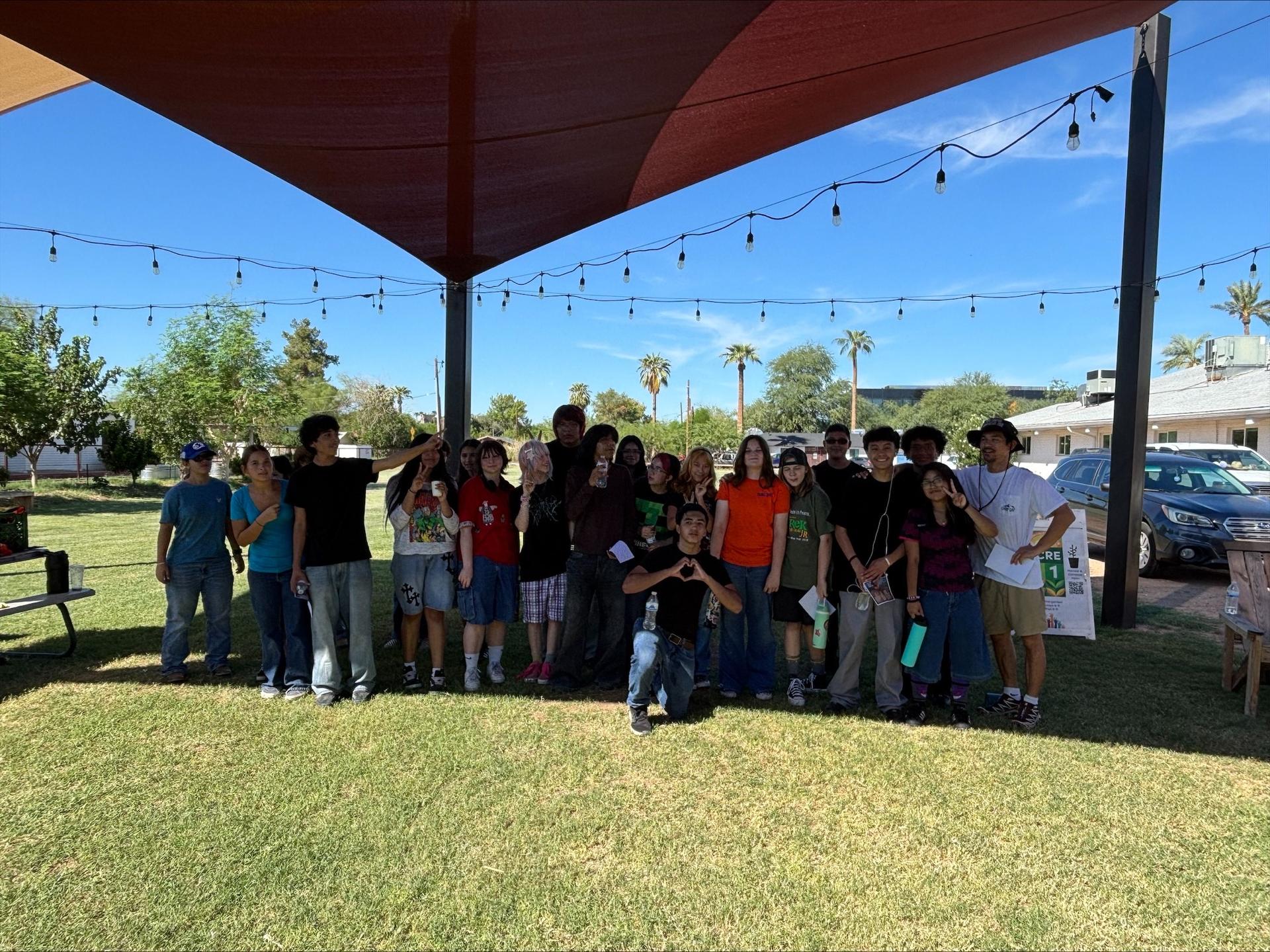 A group of young people stand together in a park under a large canopy. Some hold cameras.