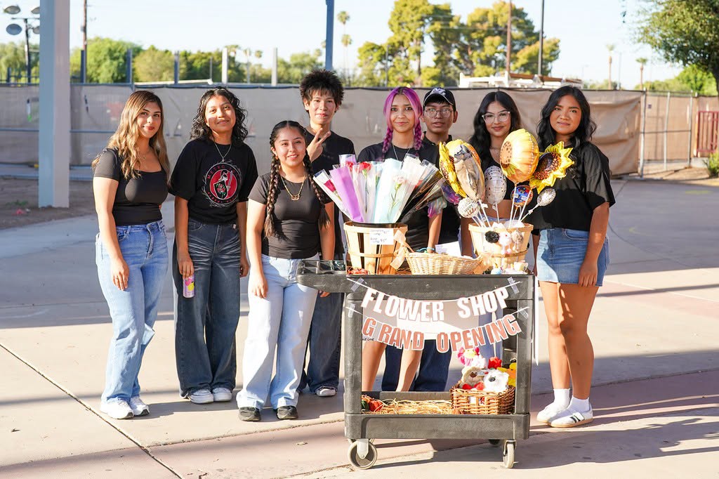 Nine young people standing near a cart with flowers. One has a balloon and a sign reading 