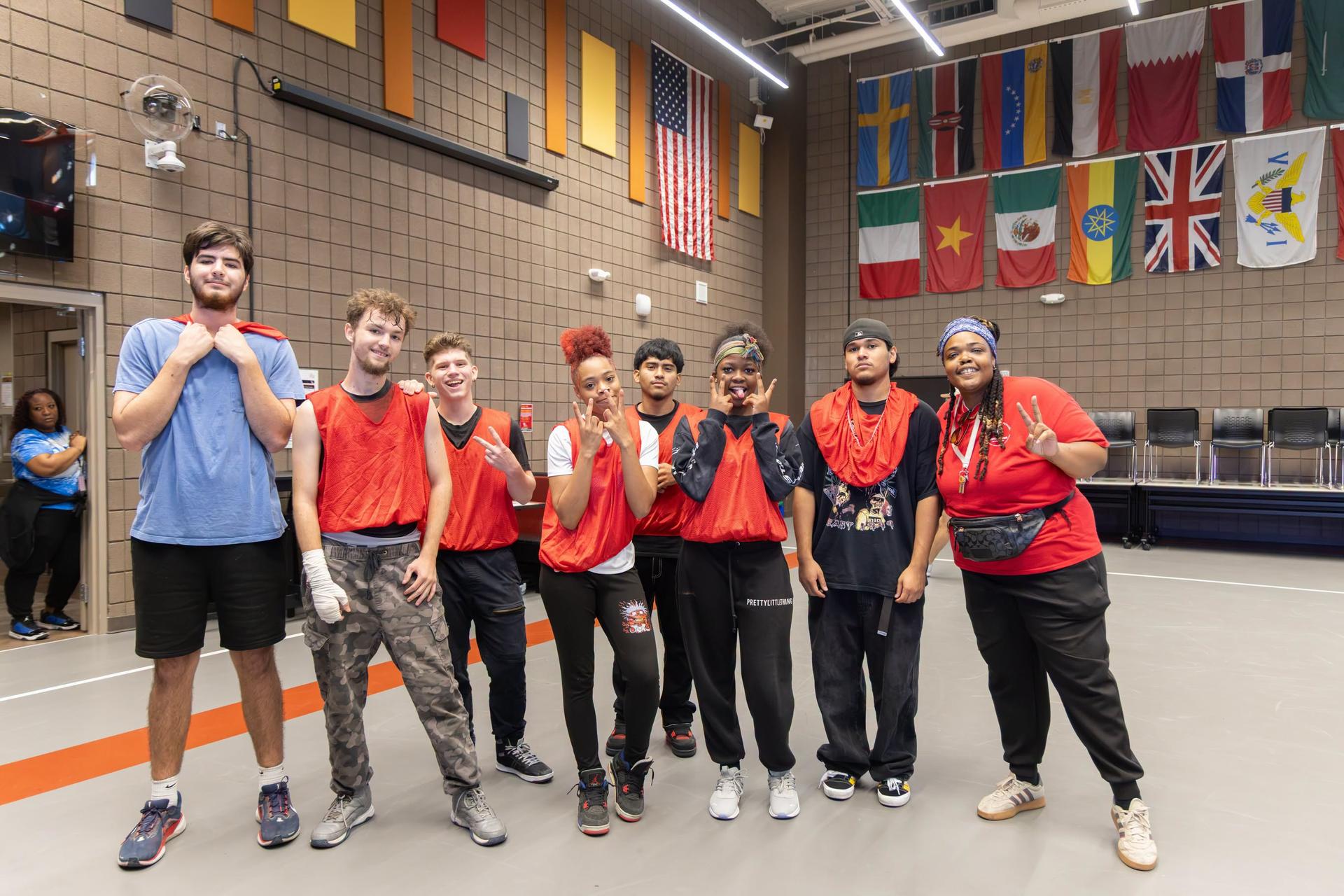 Nine people in orange shirts and black pants pose for a photo in a gym with flags.