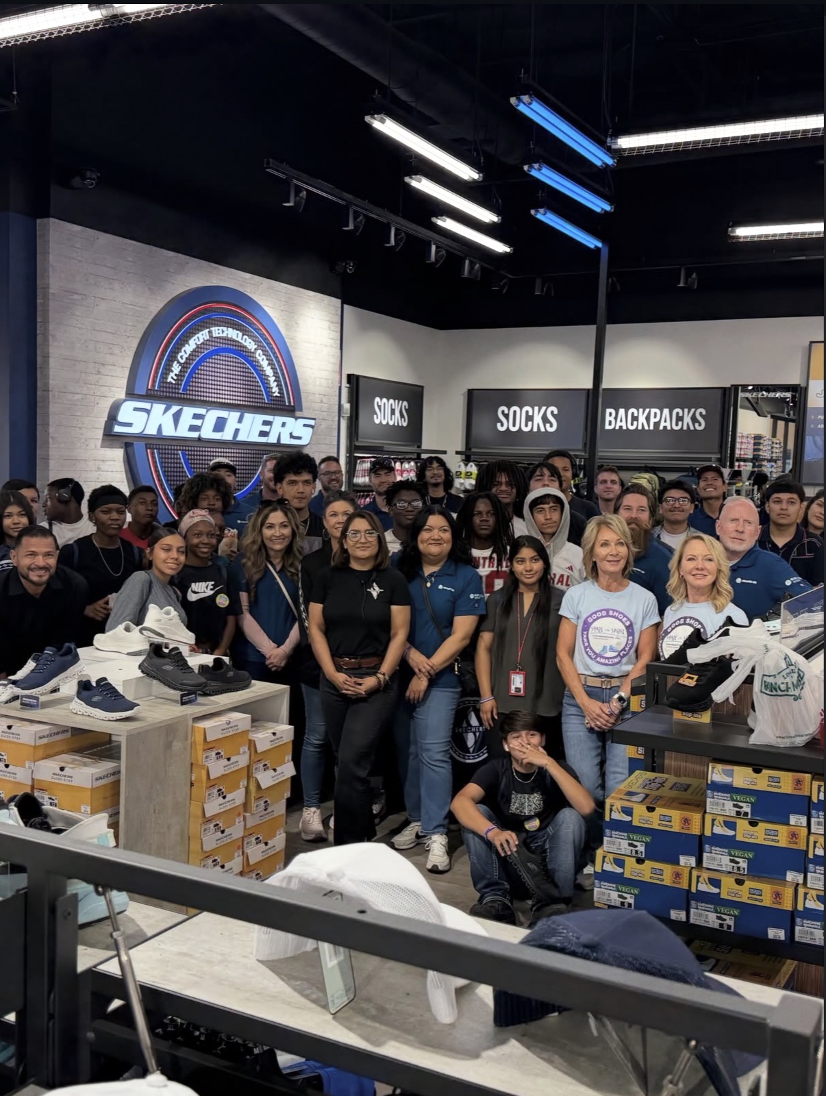 Large group of people in a shoe store. A Skechers sign is visible. Piles of shoes and boxes on shelves.