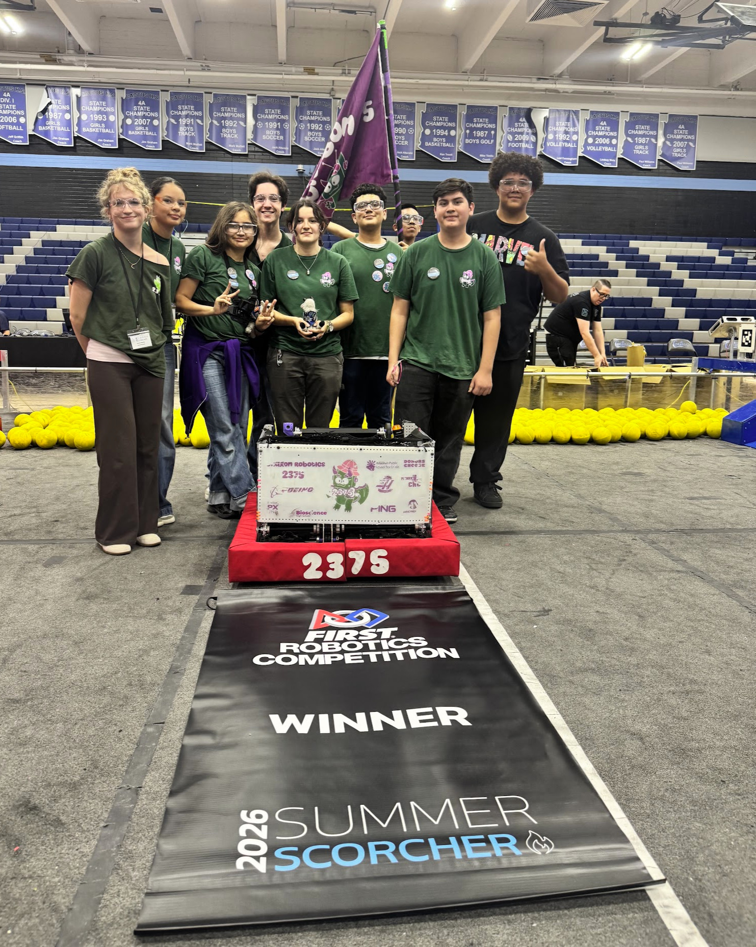 A team of students in green shirts, standing with trophies, next to a robot labeled 2375.