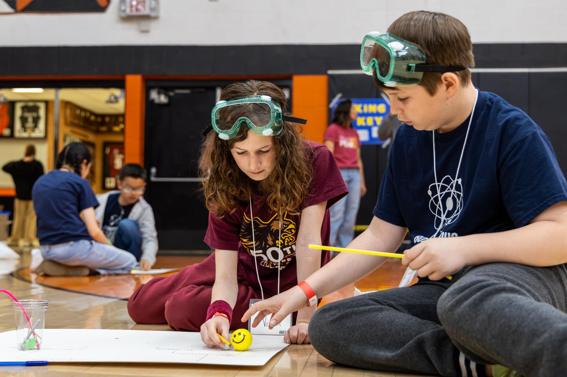 2 students holding a straw and pencil and using a ball 
