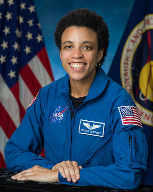 Woman in blue space suit with American flag patches. She smiles at the camera. Background includes two flags.
