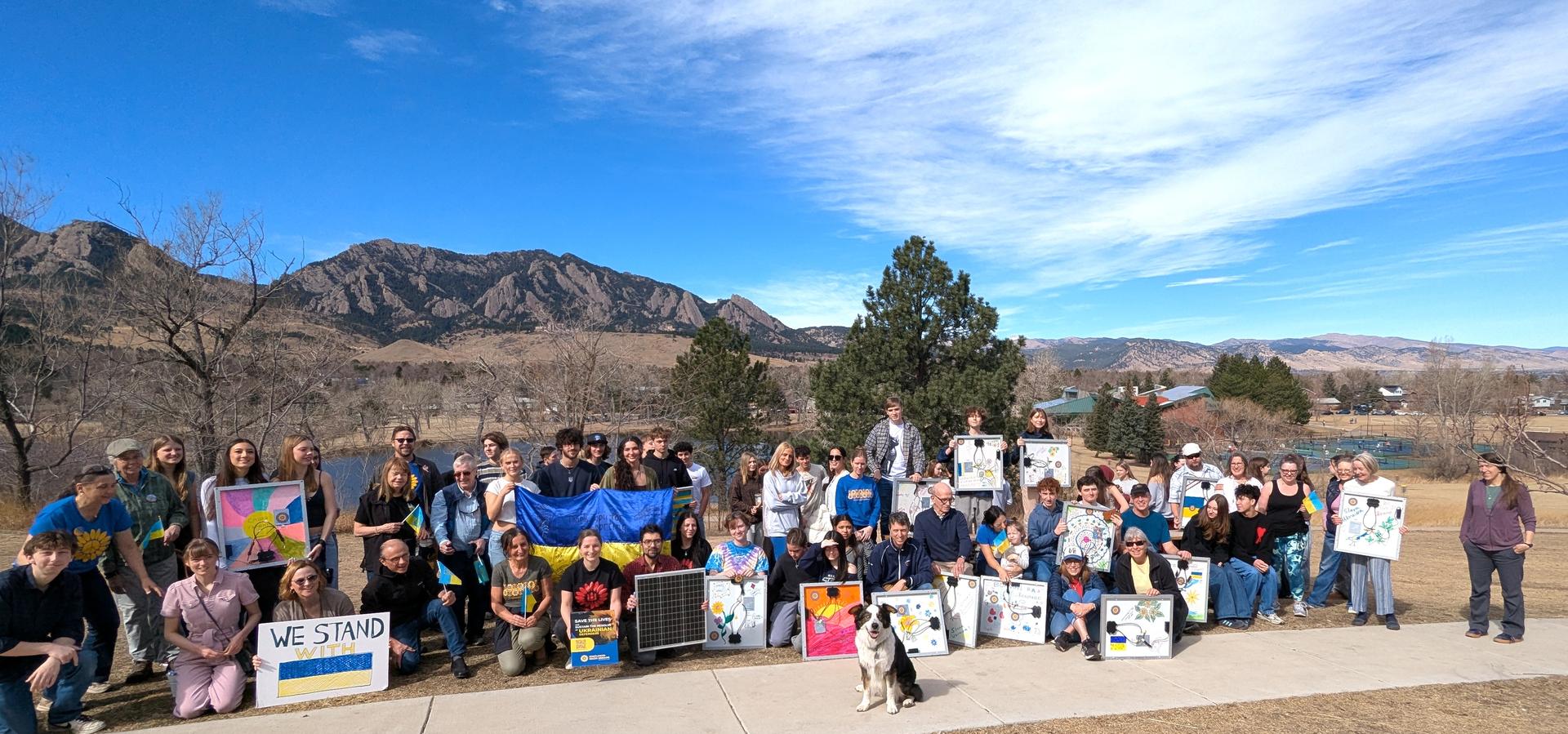 A group of people stands together holding signs. They are in front of mountains. The sky is blue with scattered clouds.