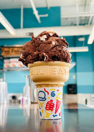 A chocolate ice cream cone with sprinkles stands on a counter. Background includes blue walls and ceiling lights.