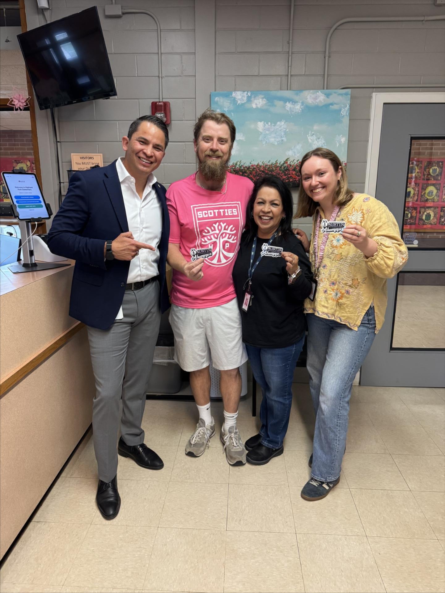 Four adults pose together indoors in a school entry area, smiling and holding up “Austin ISD Alumnus” stickers; one person wears a navy suit, another a bright pink “Scotties” shirt, with a front desk, wall-mounted screen, and a large painting in the background.