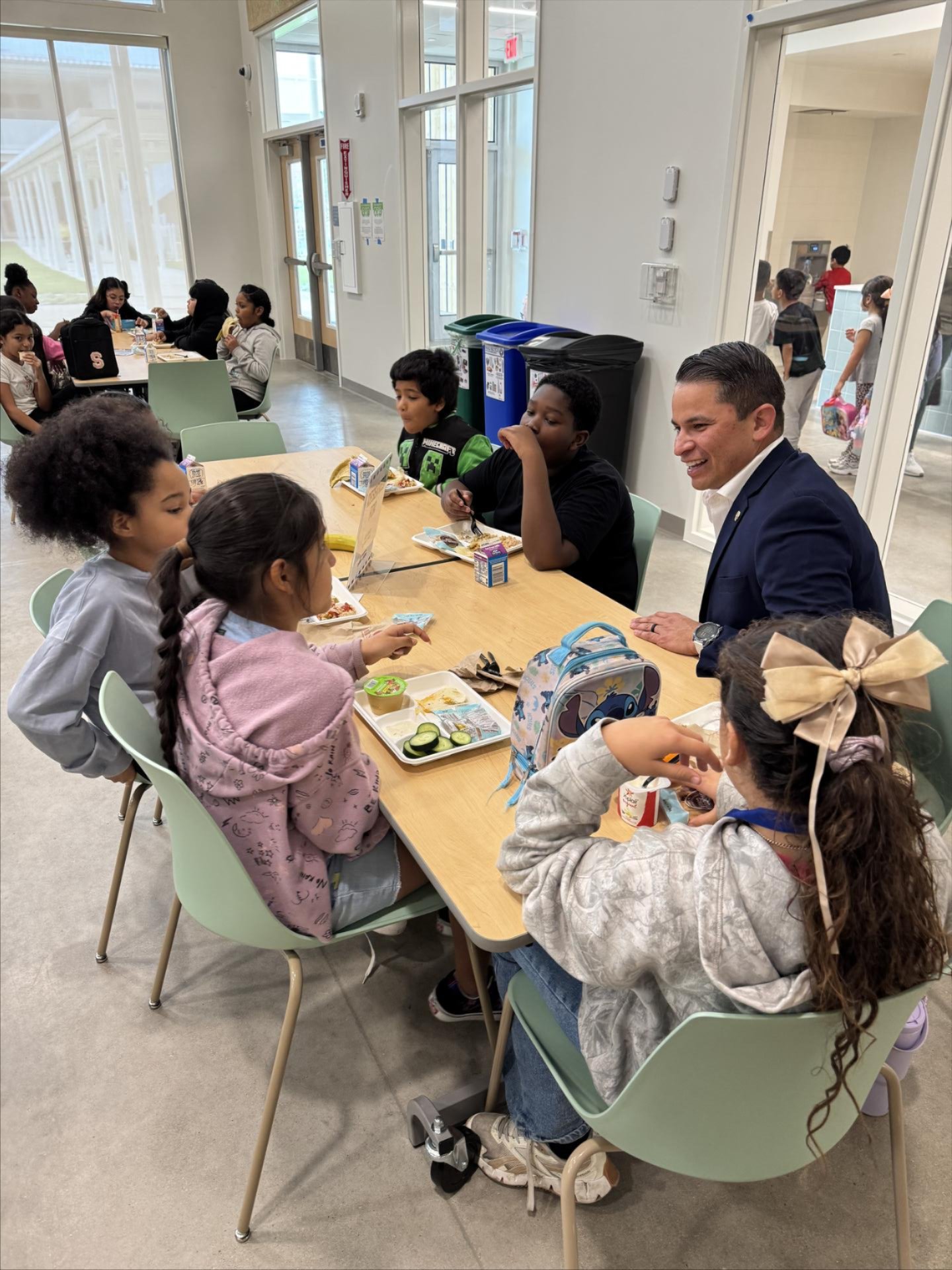 An adult in a navy suit sits at a cafeteria table talking with several elementary-aged students as they eat lunch from trays with milk cartons and snacks; other students are seated and walking in the bright, open lunch area in the background.