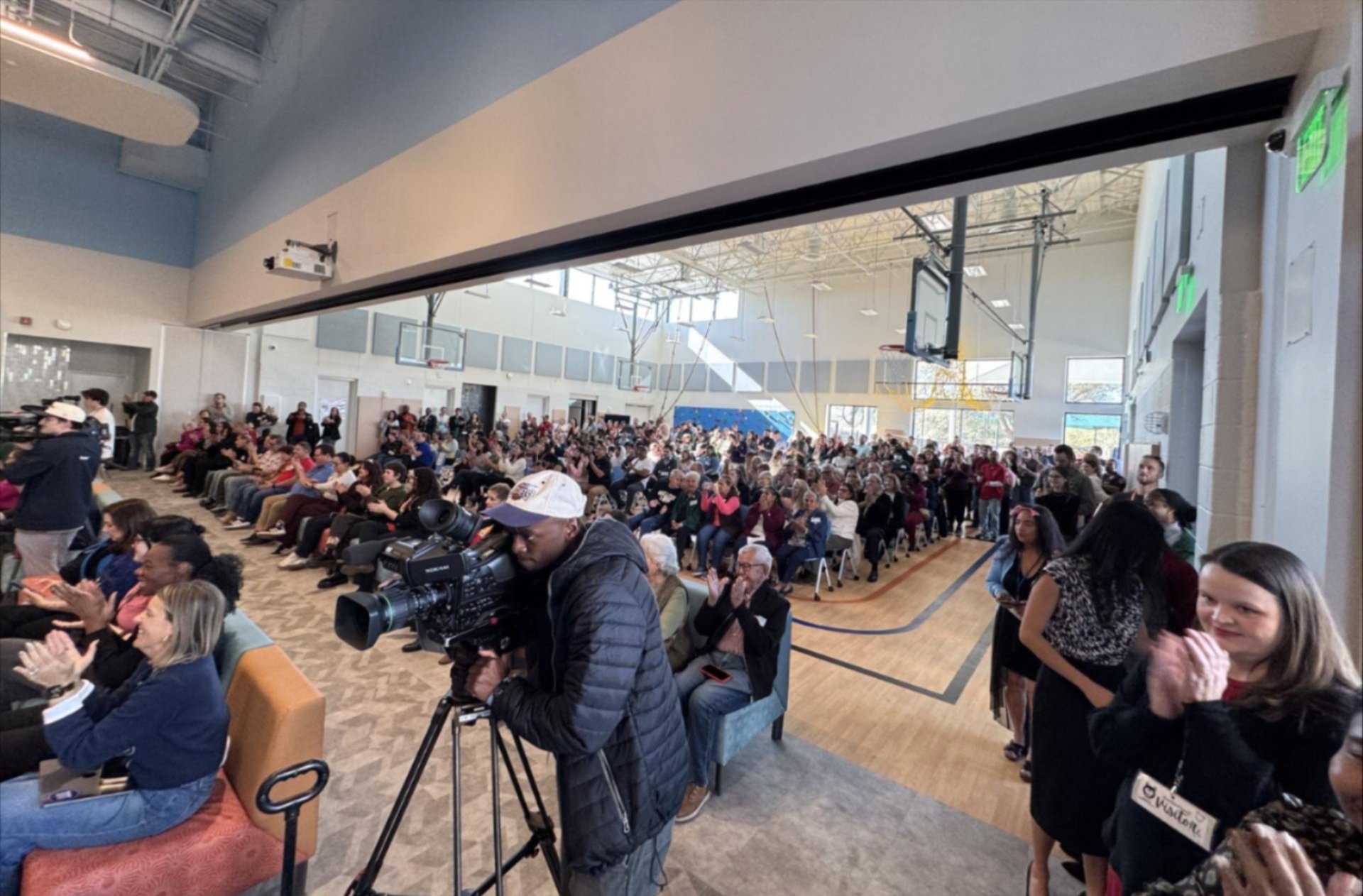 A large crowd fills a bright school gymnasium for an event, applauding as a videographer films on a tripod-mounted camera in the foreground; rows of seated attendees extend across the gym and adjoining seating area.