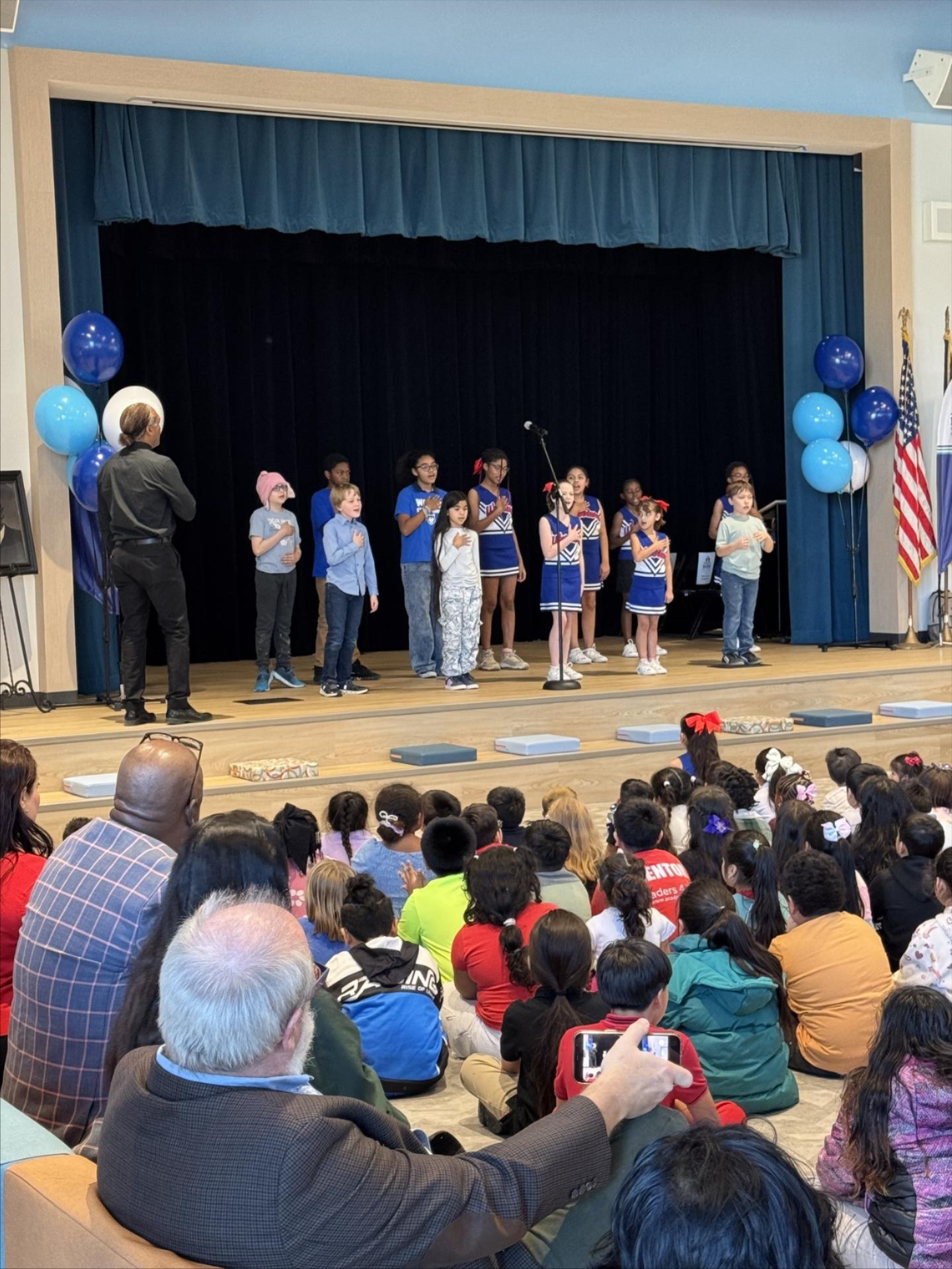 Elementary students stand on a school stage with hands over their hearts near a microphone while a large seated audience of students watches; blue and white balloons and U.S. flags are displayed at the side of the stage.