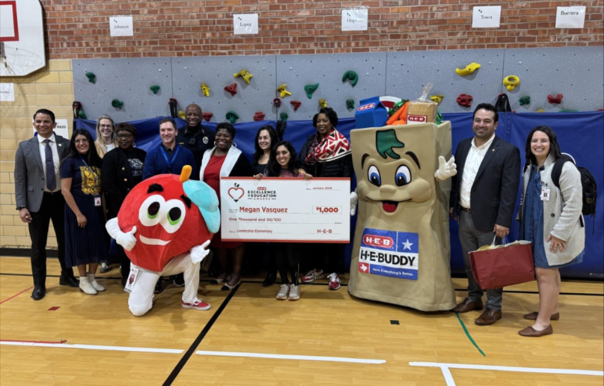 Alt text: A group of Austin ISD staff and community partners pose in a school gym with the H-E-B Buddy mascot and another mascot, holding an oversized H-E-B Excellence in Education Awards check for $1,000 made out to Megan Vasquez; a climbing wall and brick wall are in the background.