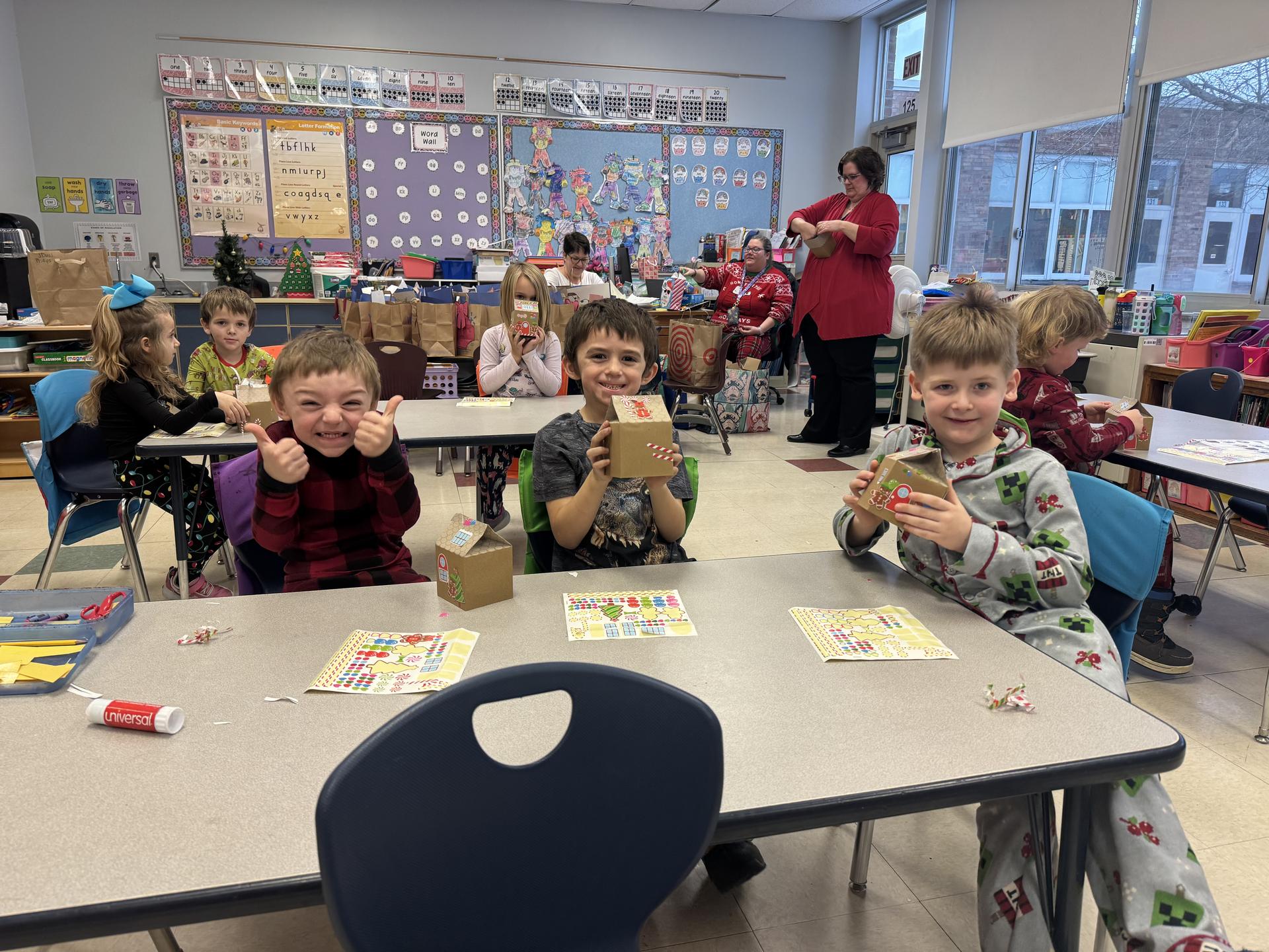 Three boys and a background of kids and a teacher smile while working on cardboard gingerbread houses