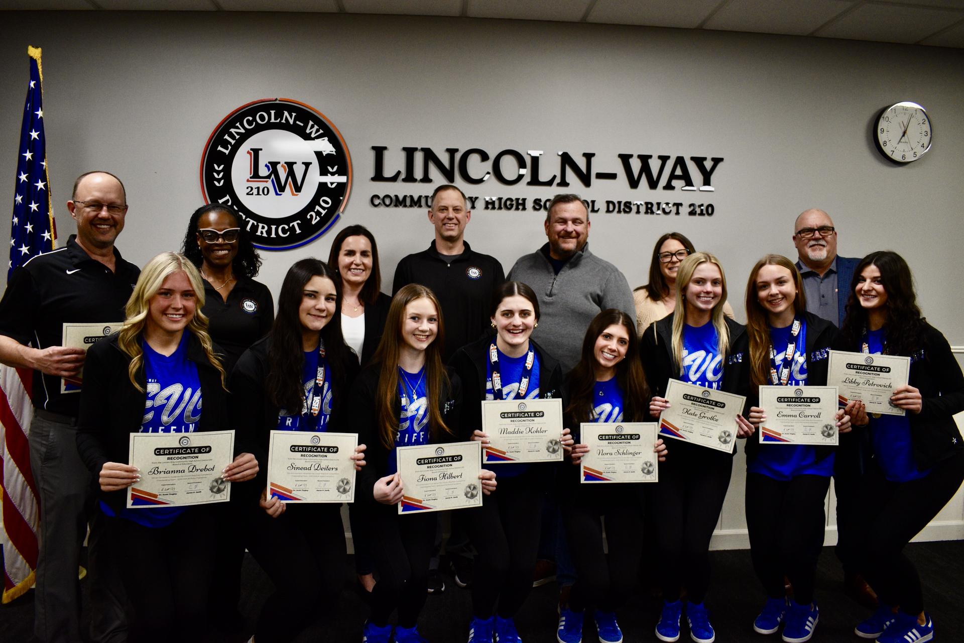 The Board of Education poses for a photo with the East competitive cheerleading team and head coach Jason Polad. A District 210 logo and an American flag are in the background.