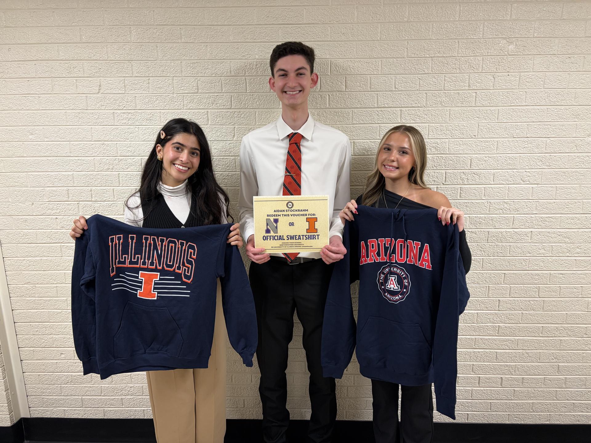 Three students stand against a blank wall for a photo, two hold a sweatshirt and one holds a certificate