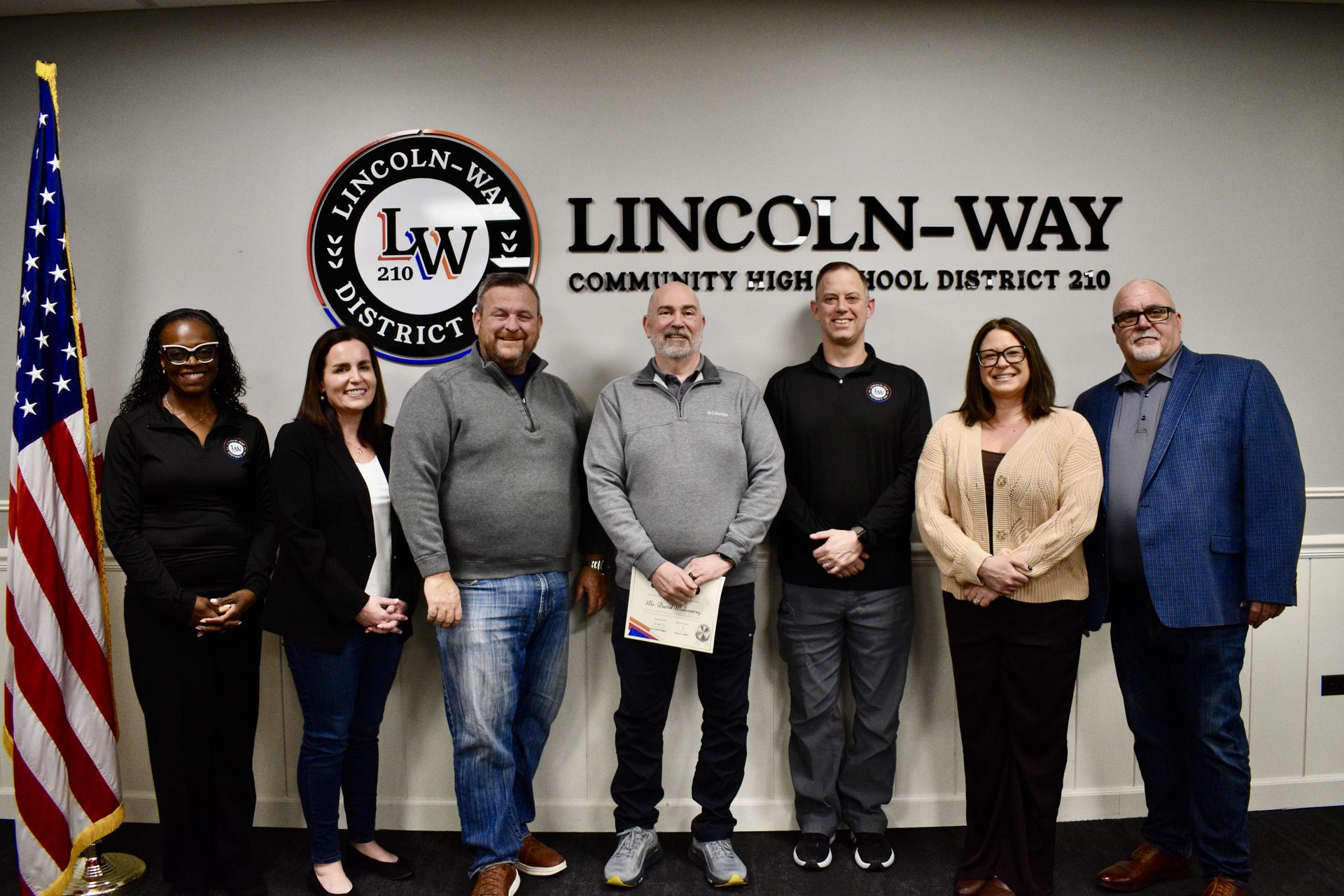 The Board of Education poses for a photo with Mr. David Mulconrey. A District 210 logo and an American flag are in the background.