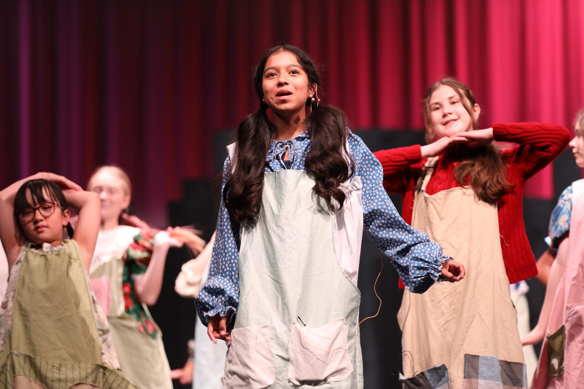 A group of elementary-aged students perform on stage in a school play, wearing simple costume outfits. A girl stands at the center singing or speaking, while other children pose and move behind her against a red curtain backdrop.