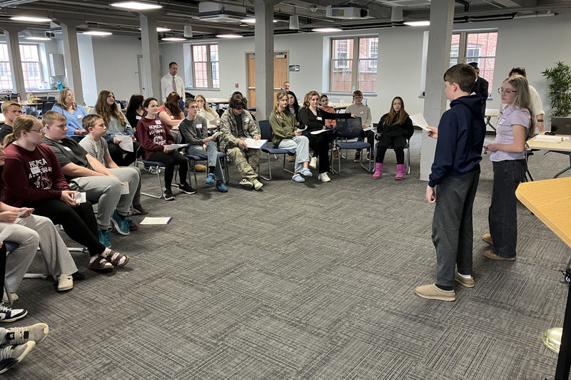 Students sitting in chairs as other students present up front