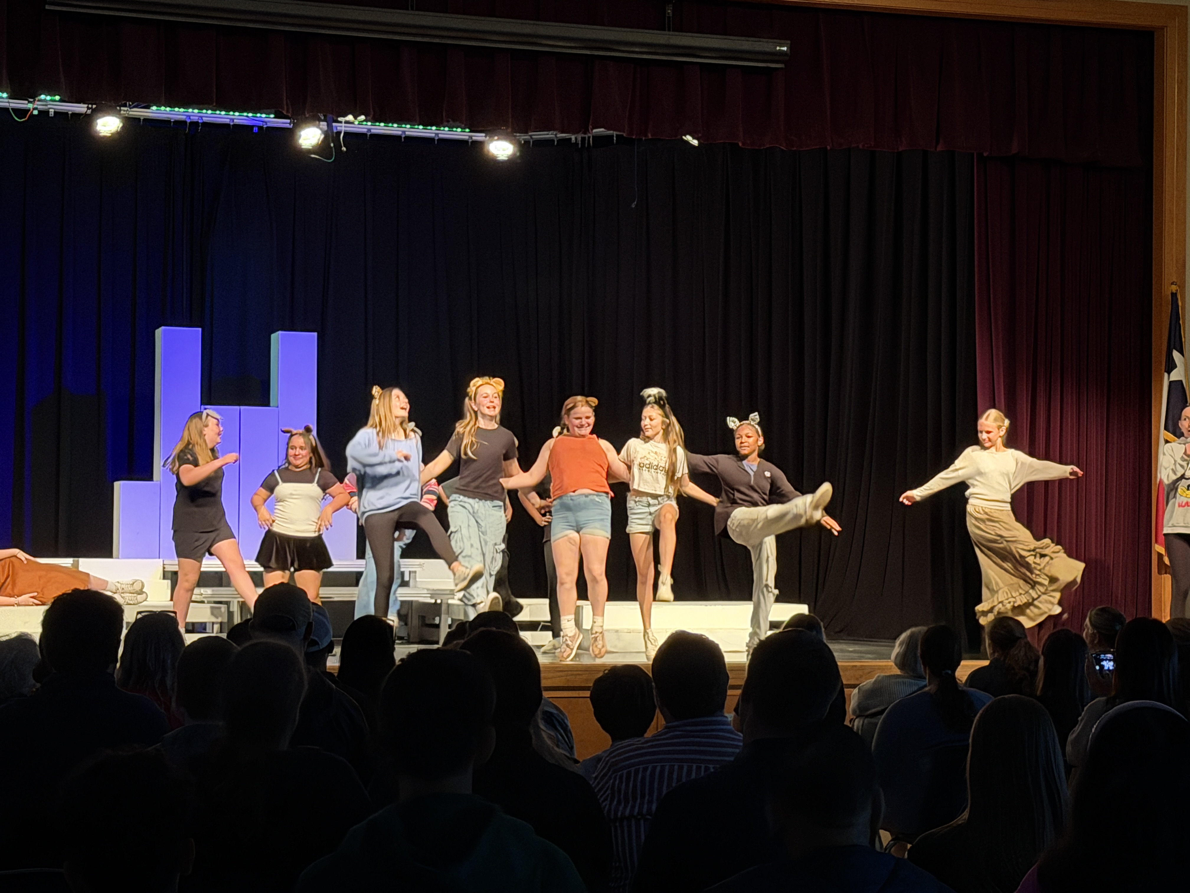 A wide-angle shot from the audience's perspective showing a group of middle school students performing on a stage during a theater showcase.