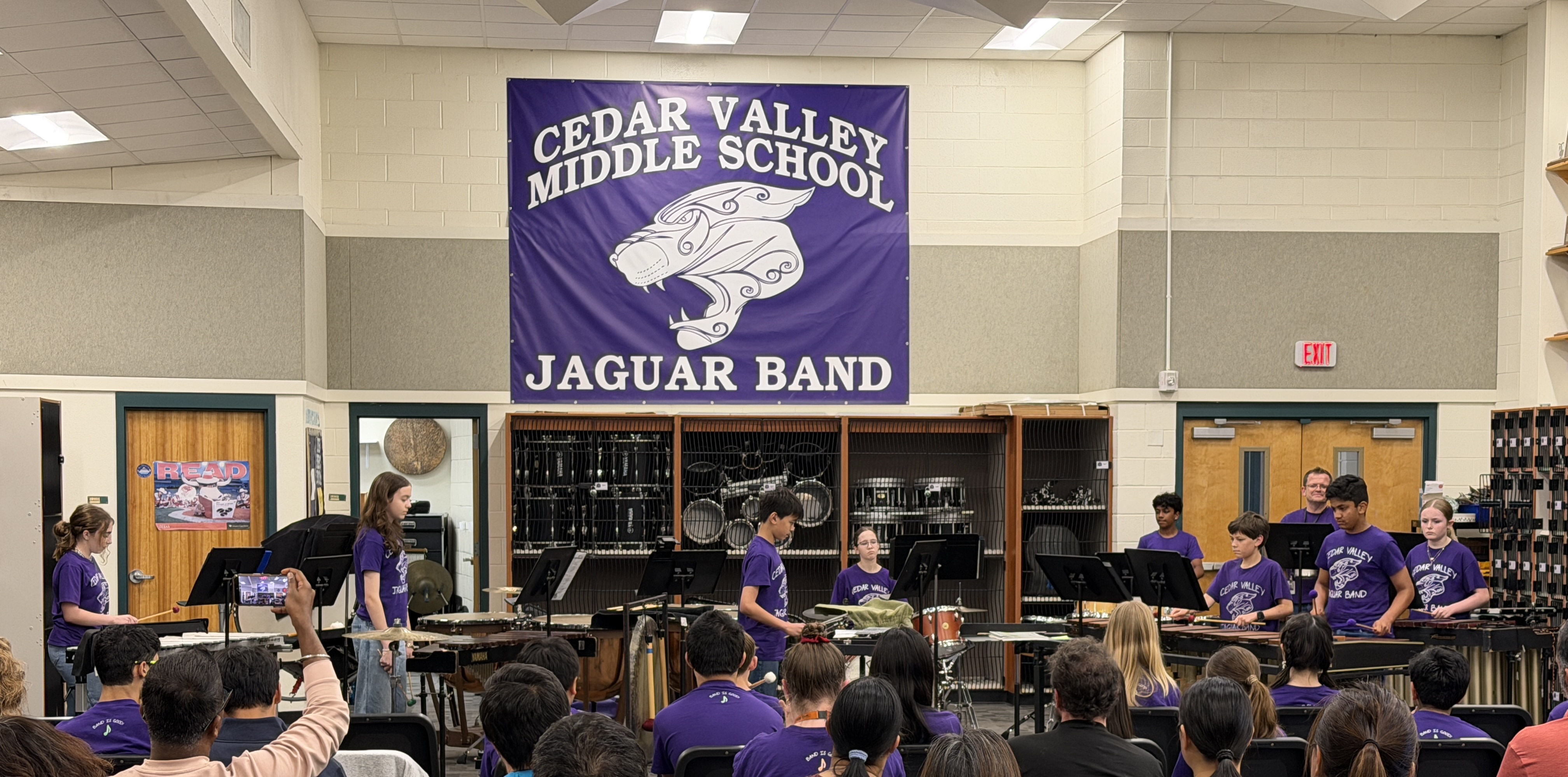A wide shot of a middle school percussion ensemble performing for an audience in a band hall.