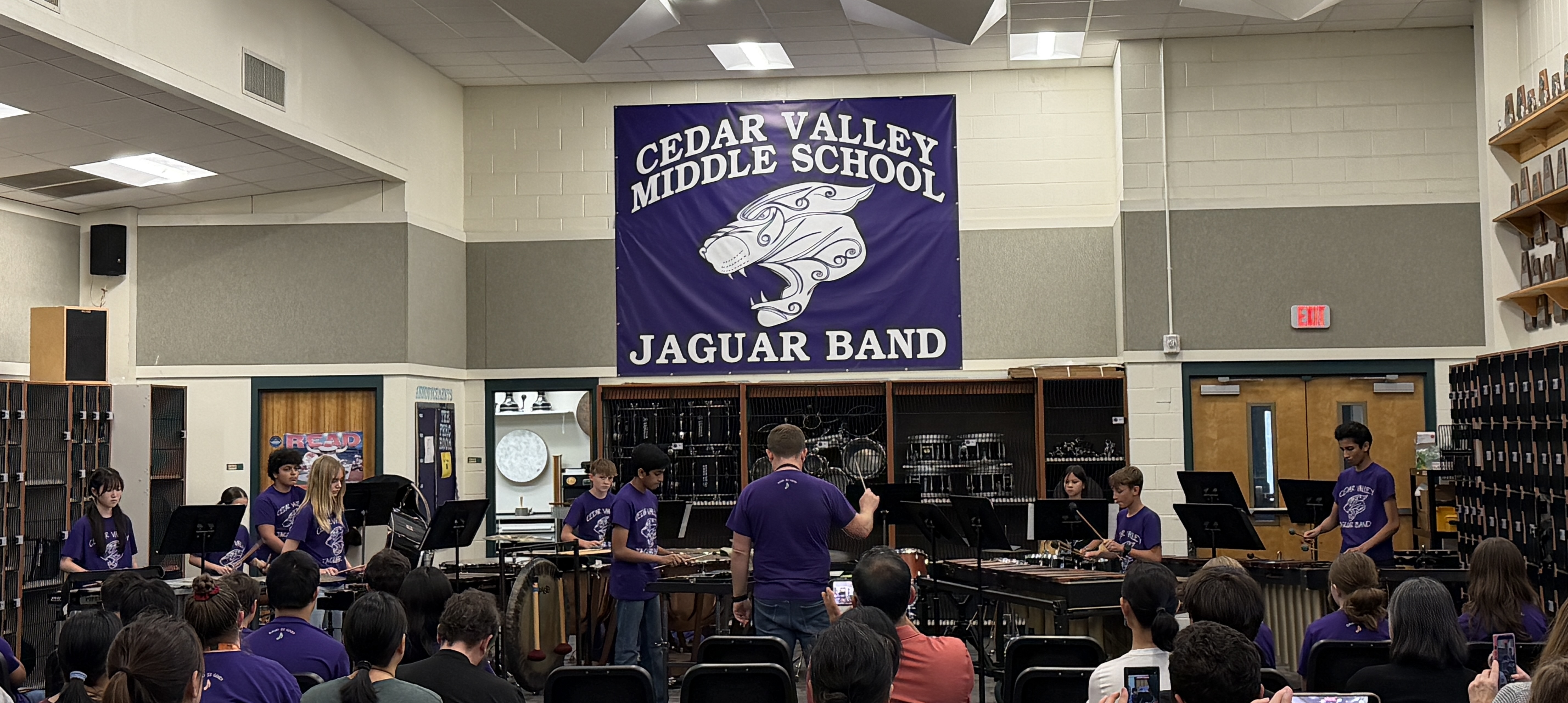 A wide shot of a middle school percussion ensemble performing for an audience in a band hall.