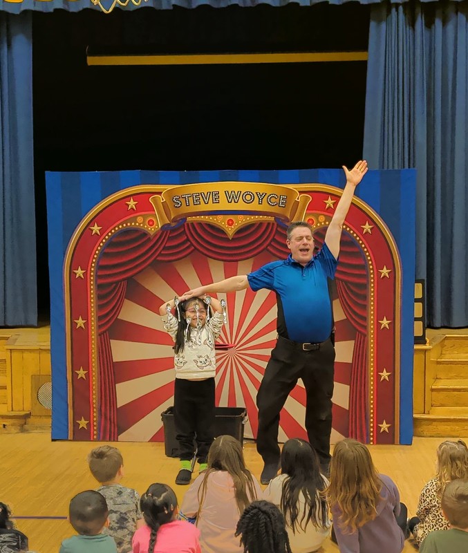 Magician Steve performs a floating spoon trick with a student in front of a group of other students in the All-Purpose Room
