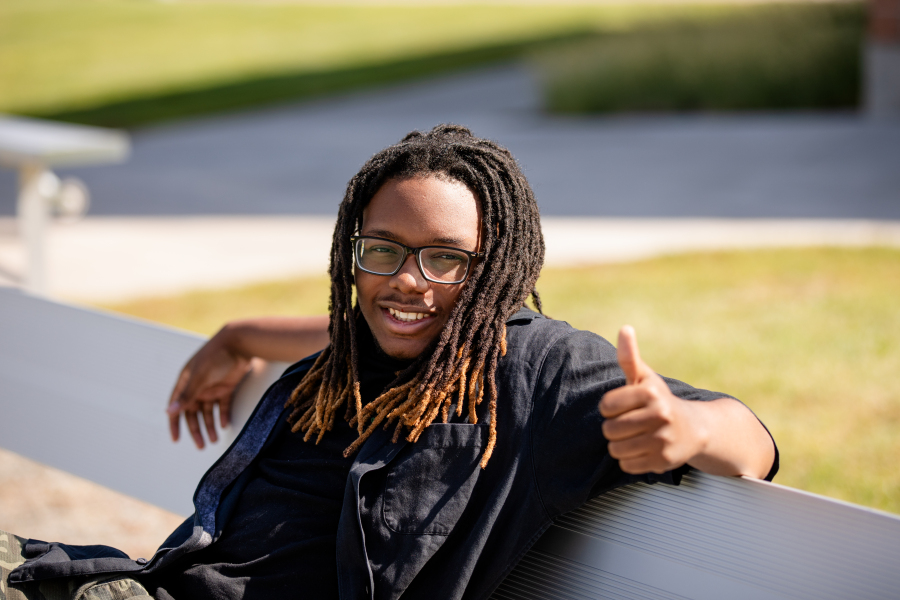 NEC Student smiles and gives a thumbs up while sitting on a bench outside.