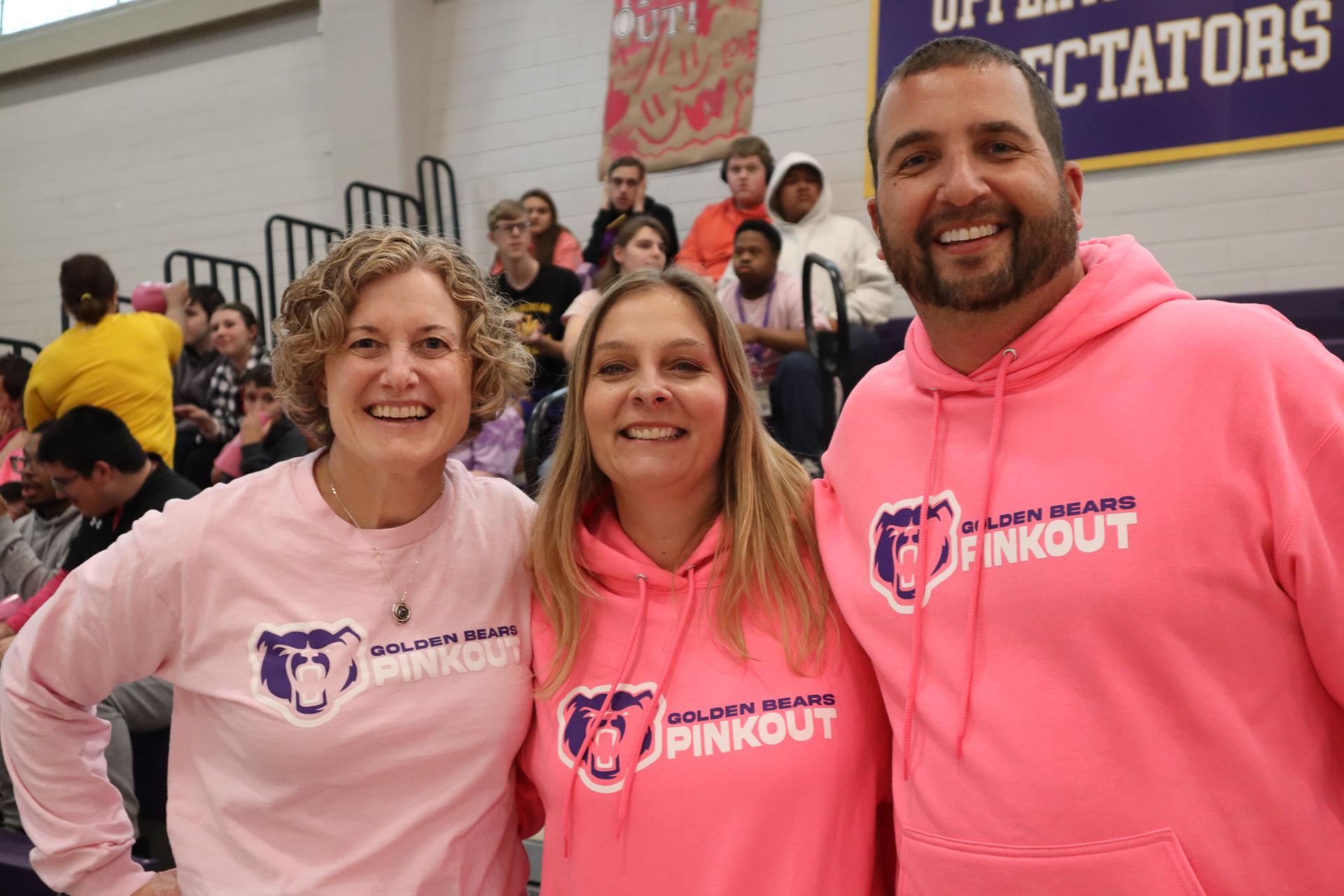 Three people smiling wearing Pink Out shirts and sweatshirts during the pep rally.