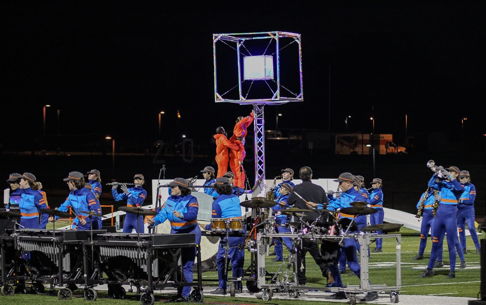 A marching band performing at night on a football field, featuring musicians in blue and orange uniforms and a central metallic cube structure with two people in orange suits climbing it.