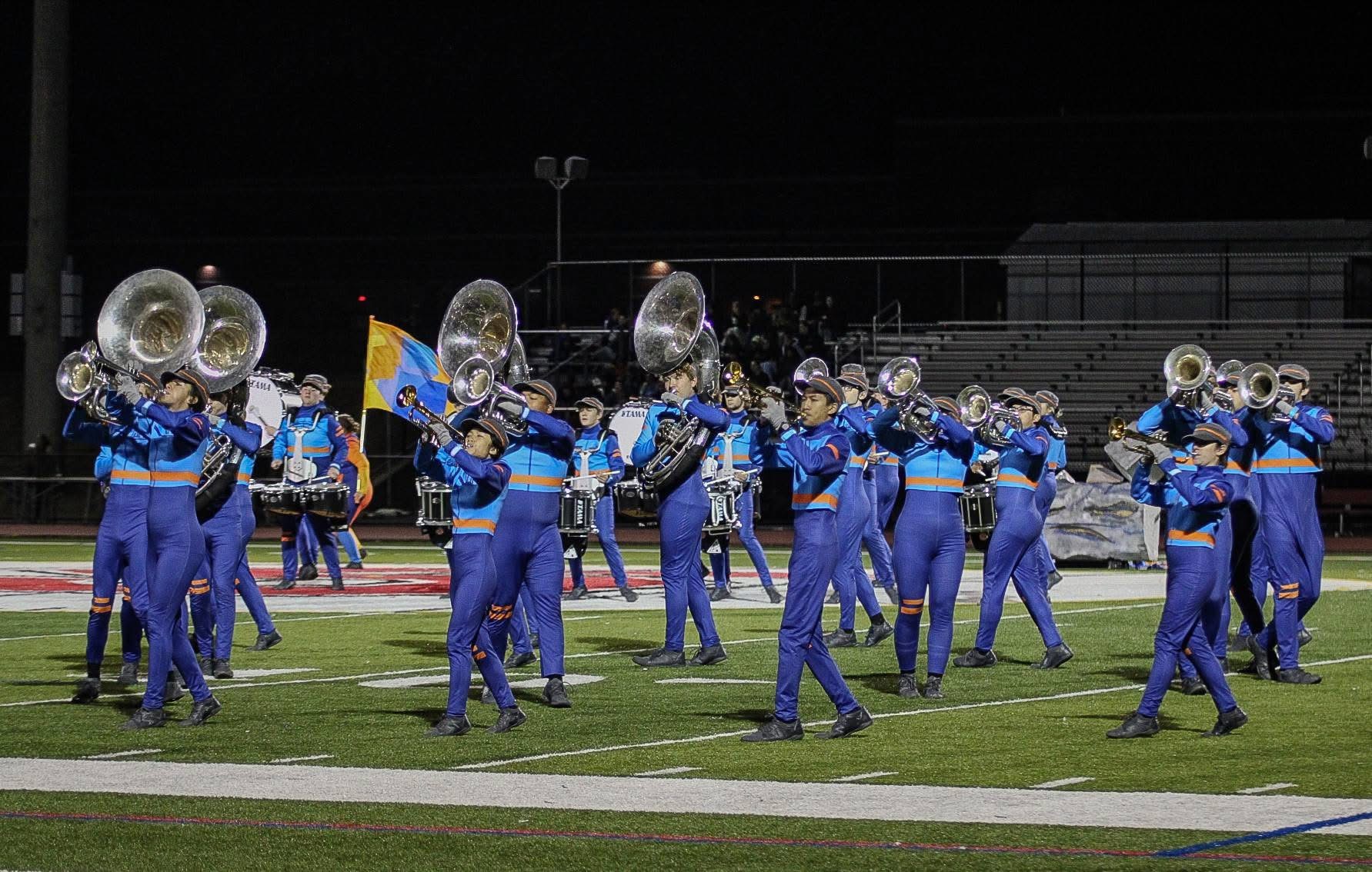 A marching band in blue uniforms with orange accents performs on a football field at night, playing brass instruments and carrying a flag.