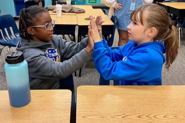 Two children sitting at a table, playing a hand-clapping game in a classroom.