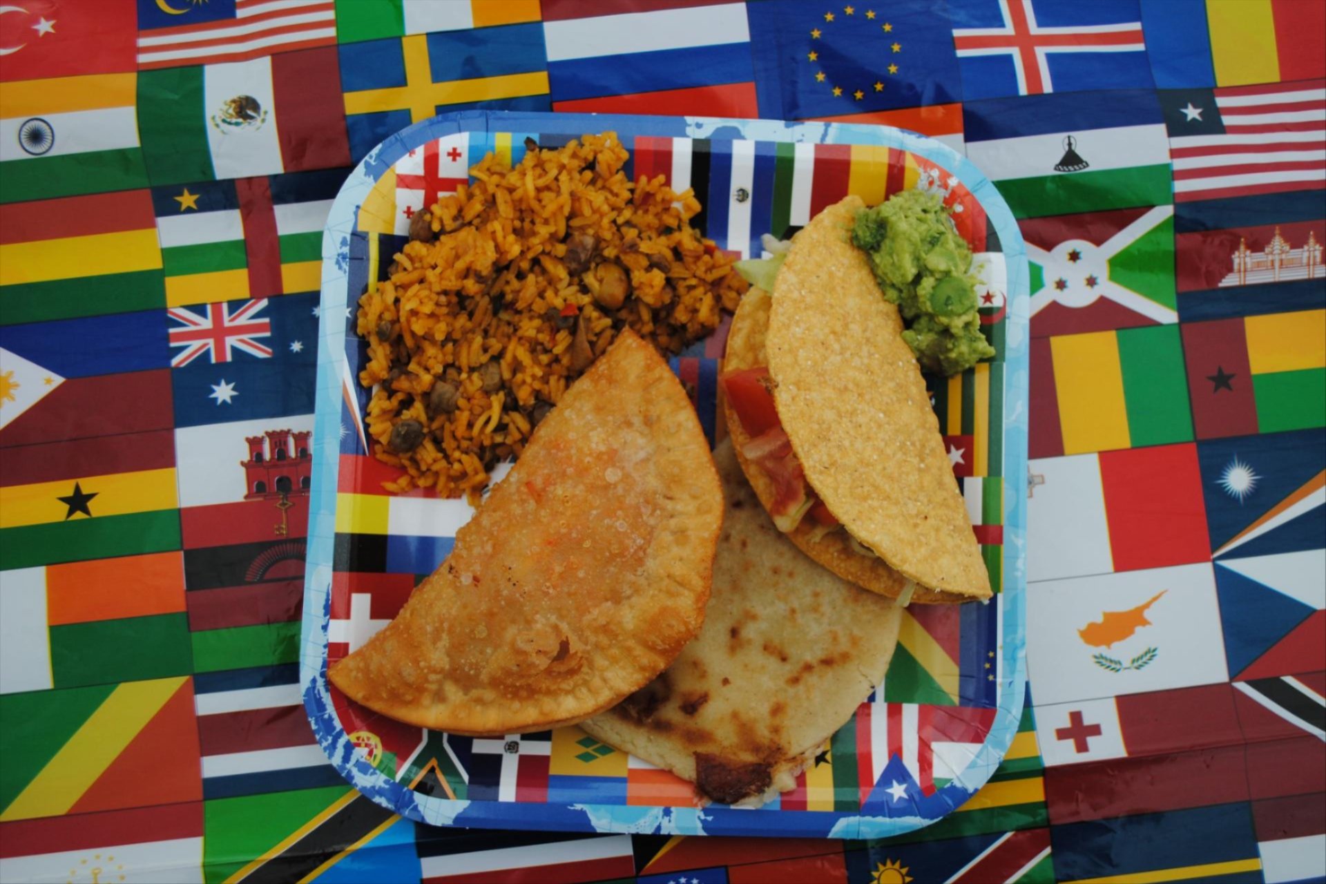 Tacos, papusas, empanadas, and rice on top of a plate and tablecloth displaying flags of the world.