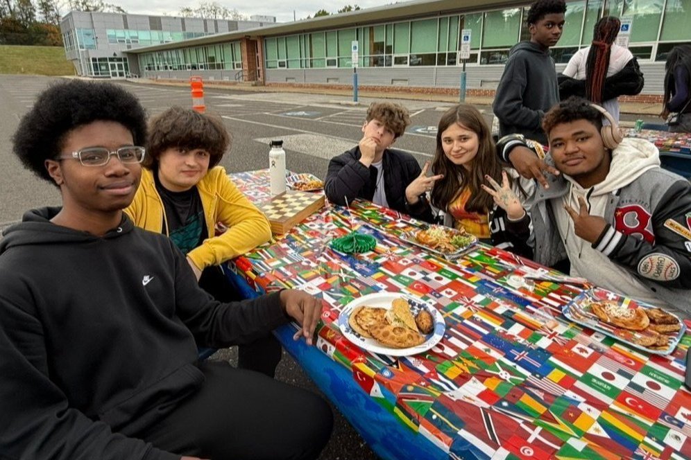 Group of students seated at a table outside, eating Hispanic foods at a table covered in a tablecloth with flags of the world. 