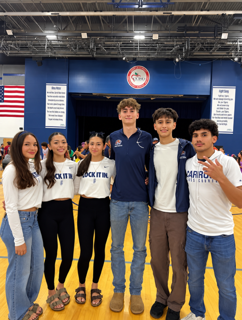 Five individuals in casual clothes, standing together in a gymnasium with a blue backdrop, smiling for a photo.