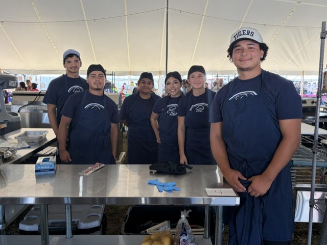 A group of people in blue uniforms and aprons stand behind a table at a food stall.
