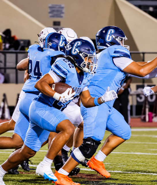 A group of football players in blue uniforms run on a field. Two players in front hold a ball.