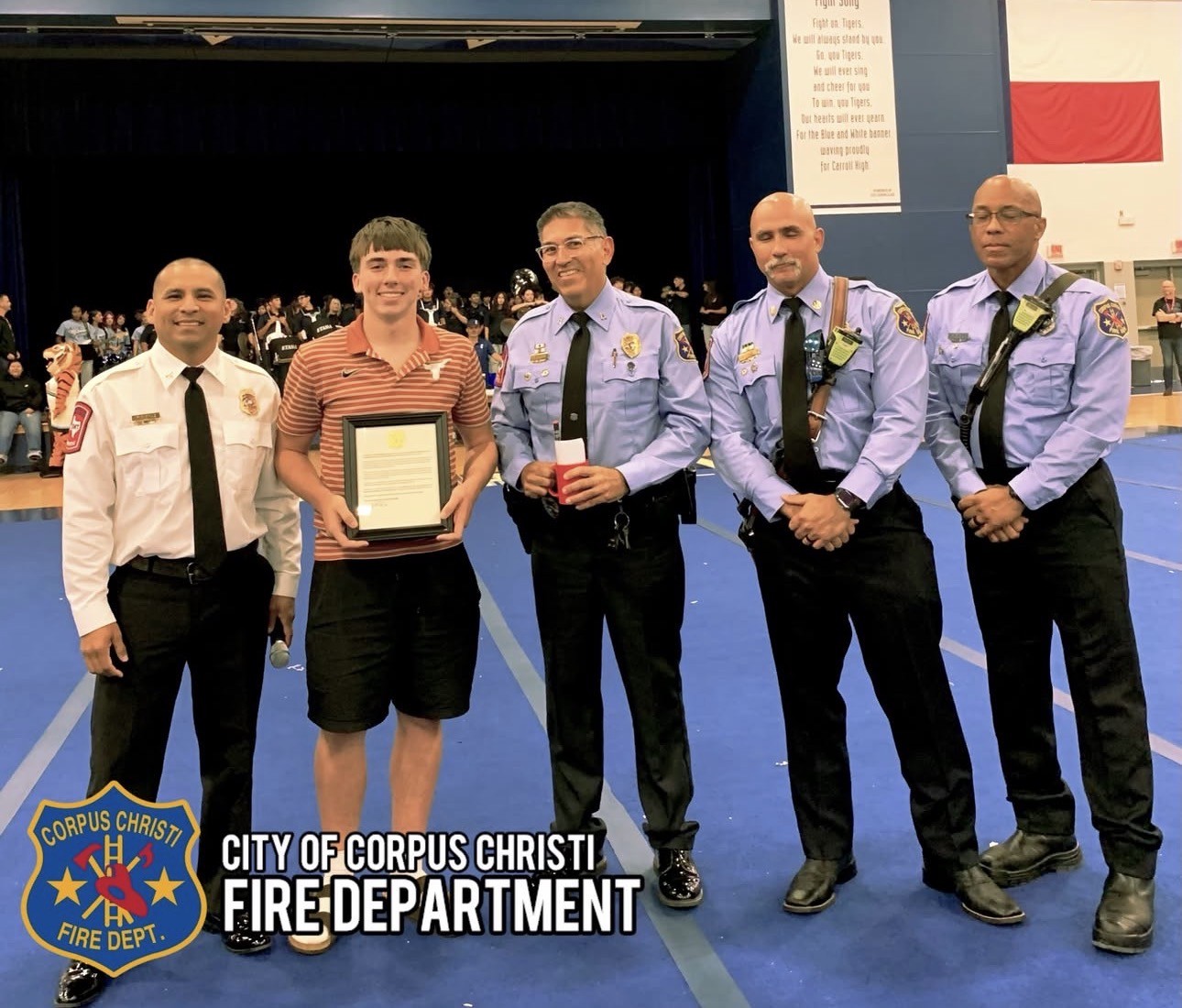 Five men stand together on a blue floor. The man in the center holds a framed award. Behind them, a banner reads 'City of Corpus Christi Fire Department'.